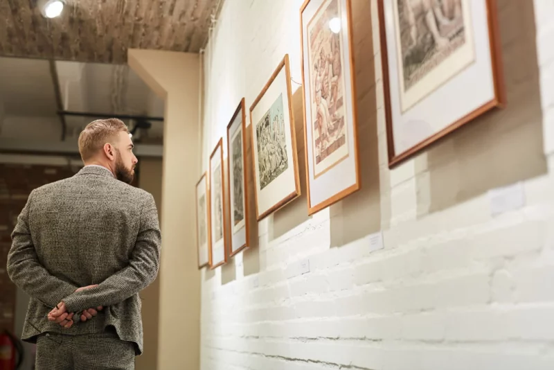 A man in a tweed suit looking at a row of framed prints on a white brick wall